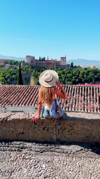 woman in straw hat sitting on rooftop overlooking alhambra palace and red tiled roofs in granada spain, vertical footage