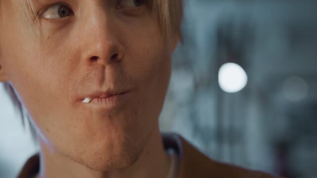 Close up of young man with short blonde hair showing subtle facial expressions and slight mouth movements. background is softly blurred indoor setting with cool lighting focusing on subject