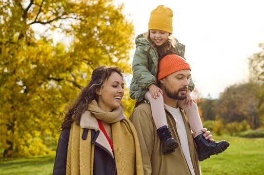 Happy family enjoys autumn walk in park. Parents in warm layers smile as their daughter rides on dads shoulders among golden leaves, sharing laughter. A warm scene of love and happiness.