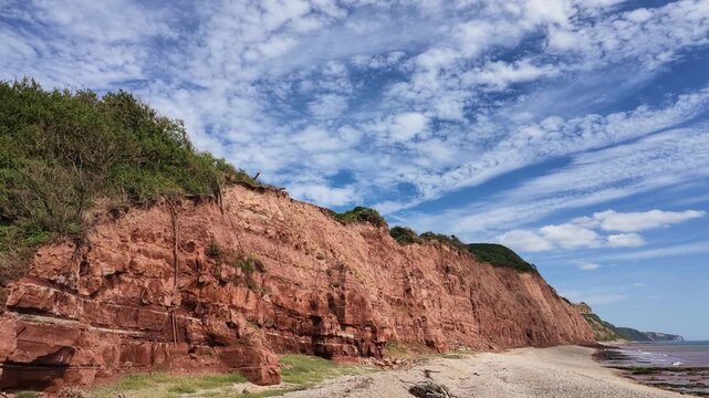 Sidmouth Devon England UK. 14.08.2026. Video. Expansive view of rugged, layered red sandstone cliffs plunging into the sea, with a sandy beach and a horizon stretching to distant headland