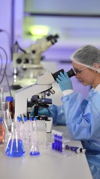 Two female research scientists in white coats, protective eyewear and sterile hairnets gowned scientist settles onto the stereo microscope as framing tightens slightly in a modern medical.