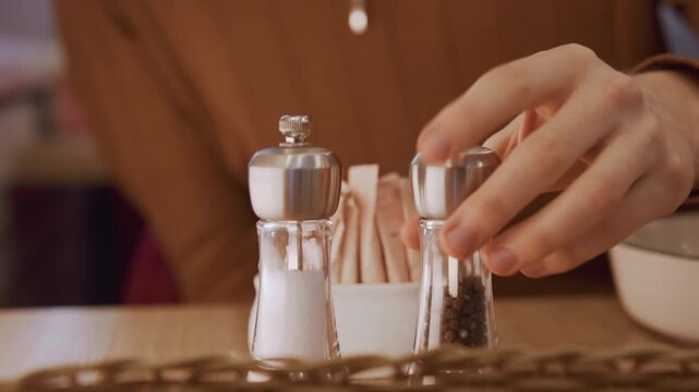 Close up of hand reaching for and placing down pepper shaker next to salt shaker on table. Background includes sugar packets woven placemat soft lighting calm expression texture detail