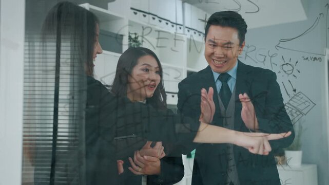 Business professionals reviewing a project plan and discussing strategy in a modern office. A female colleague points toward information on a transparent glass board while the team evaluates ideas tog