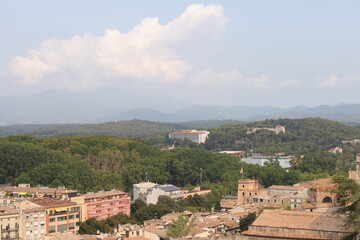 Fototapeta premium Panoramic view of Girona, Catalonia, Spain, featuring historic rooftops, lush green landscapes and distant mountain ranges under dramatic clouds. Scenic urban and natural landscape. 