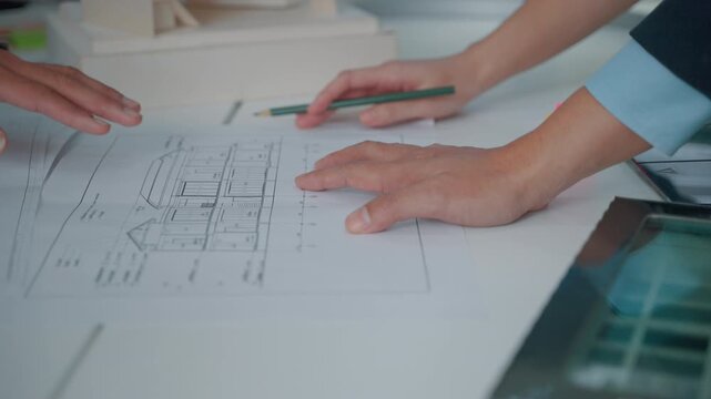Close-up of professionals reviewing an architectural blueprint and marking building plans with a pencil on a modern office desk. The image represents architecture, engineering, construction planning a