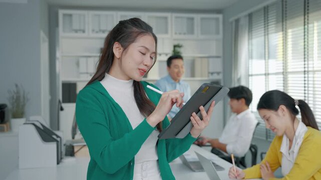 Female business professional using a digital tablet and stylus while working with colleagues in a modern office. The scene represents digital technology, workplace collaboration, productivity and prof