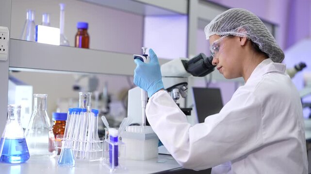 A woman researcher in a white lab coat, hairnet and clear safety goggles holding a specimen beside the microscope then reaching down to its stage in a blue-lit clinical research laboratory.