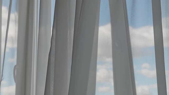 Sheer white curtains gently blowing in the wind. A bright blue sky with fluffy white clouds is visible through the translucent fabric