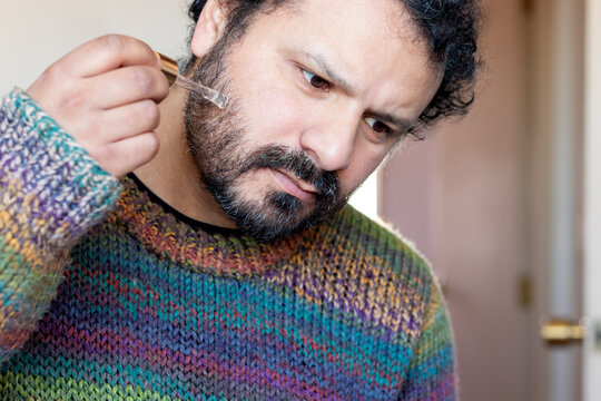 Close-up of a bearded man in a colorful knitted sweater using tweezers to pluck a gray hair from his beard at home
