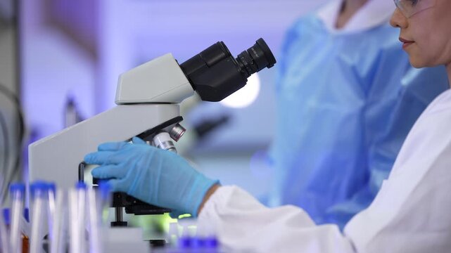 A pair of female lab technicians in white coats, protective eyewear and sterile hairnets adjusting the stage then reaching up to turn the objective turret inside a university biotechnology.