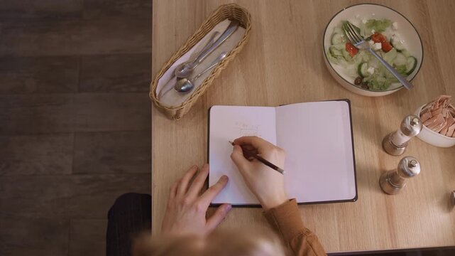 Close up of woman writing in open notebook on wooden table with soft lighting. salad bowl utensils basket and condiments are visible as woman writes continuously