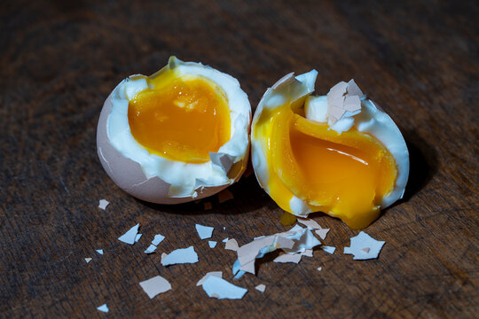 Soft boiled chicken egg cracked open into two halves showing bright runny orange yolk and white albumen on dark rustic wooden background with pieces of eggshell, closeup