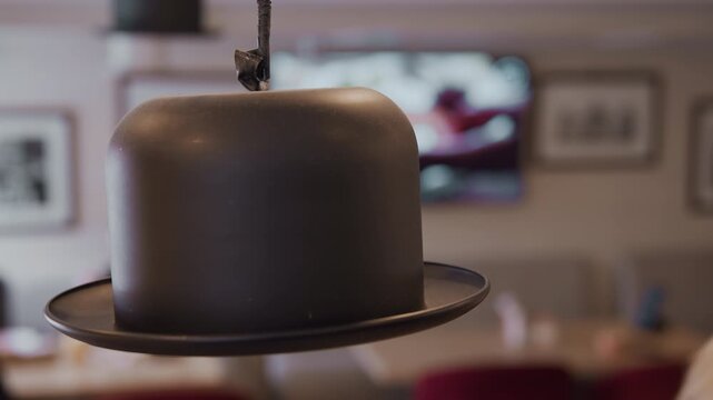 Close up of matte black bowler hat hanging from ceiling in soft indoor lighting. background features blurred framed artwork on light colored walls and indistinct figures seated at tables