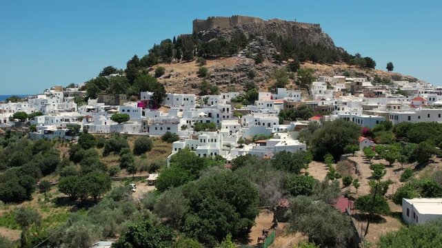Stunning aerial drone footage flying over the ancient Acropolis of Lindos in Rhodes, Greece. The footage reveals the historic ruins perched above the picturesque whitewashed village of Lindos