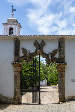 View of the historic Knots Gate with its intricate Manueline rope carvings and iron gate, set against a white wall and bell tower in Vila Vicosa, Distrito de Evora, Portugal.