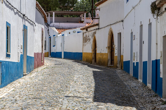 View of a narrow cobblestone street lined with traditional white-washed houses featuring blue and yellow painted bases in Vila Vicosa, Distrito de Evora, Portugal.