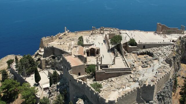 Stunning aerial drone footage flying over the ancient Acropolis of Lindos in Rhodes, Greece. The footage reveals the historic ruins perched above the picturesque whitewashed village of Lindos