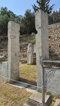 Athens, Greece - 4 July 2026. Tall marble door jambs frame an entrance beside the ancient fountain in the Roman Forum, with masonry remains and trees behind.