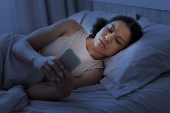 Worried young Black woman lying awake in bed at night checking her smartphone, suggesting insomnia, late-night scrolling, anxiety, notifications, or troubling online information