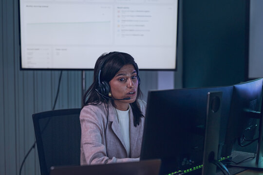 Asian female customer service professional wearing a headset works at multiple computer monitors, providing technical support with a business analytics dashboard displayed behind her