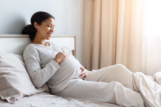 Smiling pregnant Black woman relaxes on a bed and gently holds her baby bump, expressing comfort, maternal bonding, happiness, and anticipation during pregnancy at home
