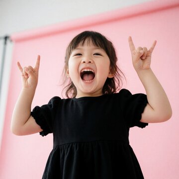 Young girl with open mouth makes rock hand sign. Asian child expresses joy and energy on pink background. Kid plays, has fun and celebrates, showing rock n roll symbol.