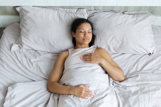 Young Black woman sleeps peacefully on her back under soft bedding, illustrating healthy sleep, rest, relaxation, recovery, and a calm bedroom routine at home