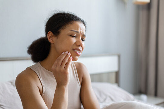 Young Black woman sits in her bedroom touching her jaw with painful expression, experiencing toothache, dental discomfort, sensitivity, or oral health problems at home