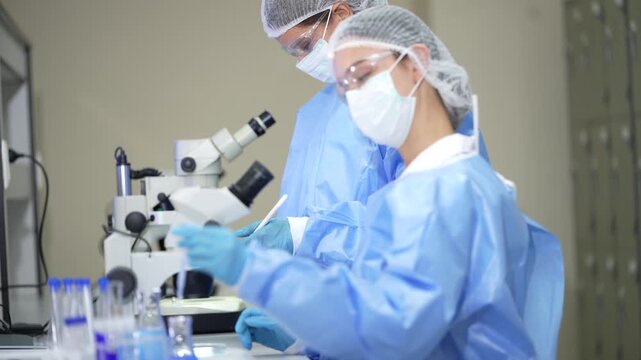 A pair of female lab technicians wearing face masks, hairnets and safety goggles gowned pair bends over stereo microscope, then lifts a swab for inspection inside a clean pharmaceutical.