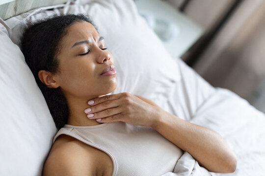 Young Black woman resting in bed and touching her throat with painful expression, experiencing soreness, illness, cold, flu symptoms, or difficulty swallowing at home