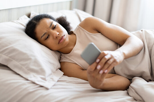 Young Black woman lies in bed looking concerned while checking her smartphone, reacting to troubling information, message, notification, or unexpected online news at home