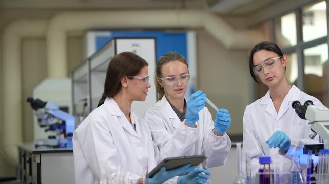 Three women laboratory colleagues in white coats, protective eyewear and sterile hairnets inside a university biotechnology laboratory, with trio talking, one lifts a sample tube, third.