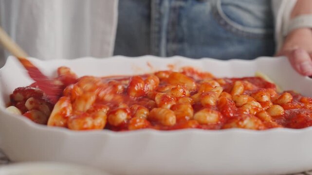 Woman mixes gnocchi with tomato sauce in white ceramic baking dish using spatula. Home cooking and homemade meal preparation