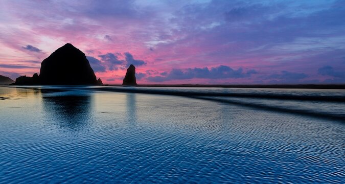 View of Haystack Rock at sunset with vibrant purple and pink clouds reflecting on the wet sand of the beach in Cannon Beach, Oregon, United States.