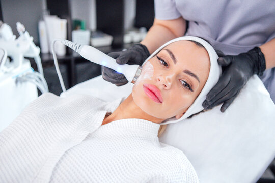 Woman receiving facial treatment in a modern spa, with a skincare specialist applying a device on her face, showcasing relaxation and beauty care in a serene environment