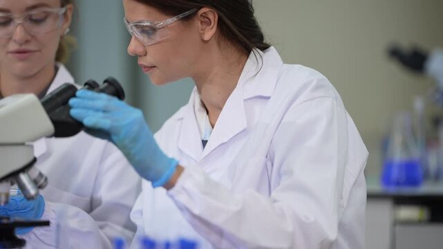 Two women scientists in white coats, protective eyewear and sterile hairnets straightening from the eyepiece and turning to an approaching colleague at a professional laboratory workstation.