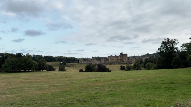 Wide landscape view of a majestic historic medieval castle surrounded by green park fields and forest trees under cloudy sky. English countryside tourism.