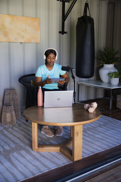 Adult African woman recording into mic holding tablet with headphones by punching bag in studio