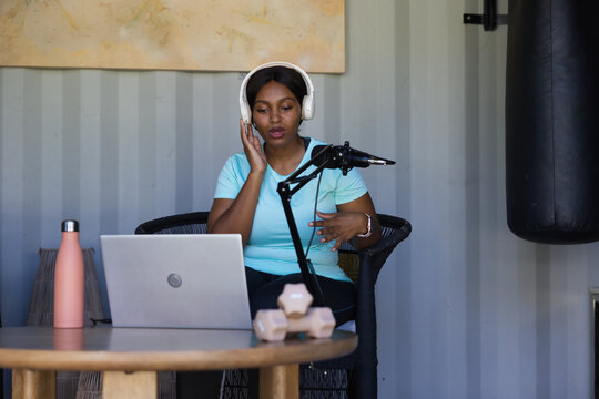 African American woman wearing white headset speaking into boom mic in studio at table with laptop