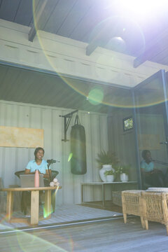 African American woman sitting in bright studio recording with laptop, mic and punching bag behind