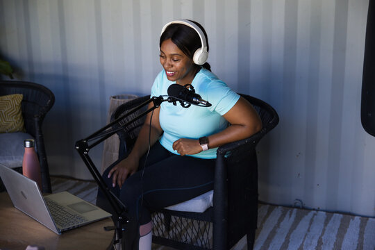 Adult African woman sitting in compact studio recording on laptop with microphone and headphones