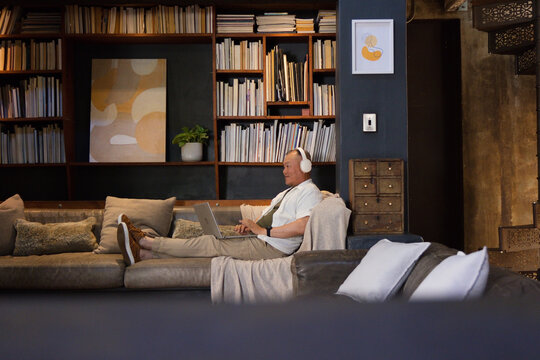 Senior Asian man reclining on sectional sofa in lounge using laptop wearing headphones, copy space