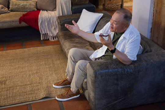 Senior Asian man sitting on sofa at home holding phone, gesturing, wearing smartwatch, copy space