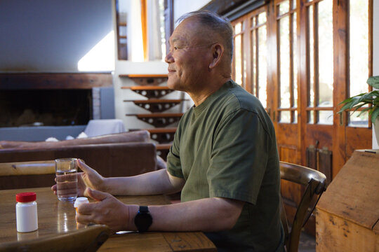 Senior Asian man at home dining table, holding water and pill bottle, wearing olive tee, watch