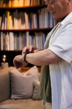 Senior Asian man standing in study by bookshelf wearing tee and adjusting smartwatch, copy space