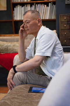 Senior East Asian man sitting on sofa leaning forward, hand on face, tablet and bookshelf nearby