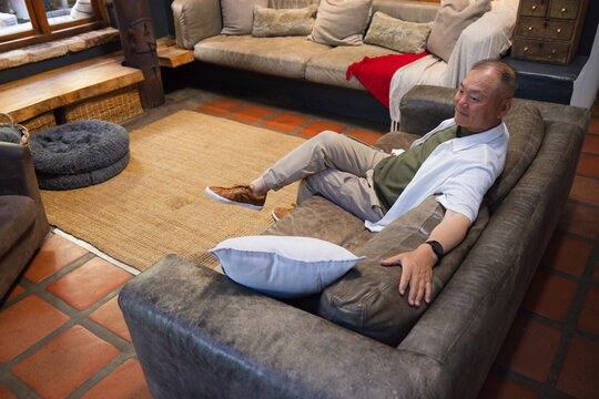 Senior Asian man reclining on grey suede sofa in living room wearing open shirt, copy space