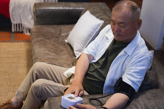 Senior Asian man sitting on leather sofa at home measuring blood pressure with digital cuff monitor