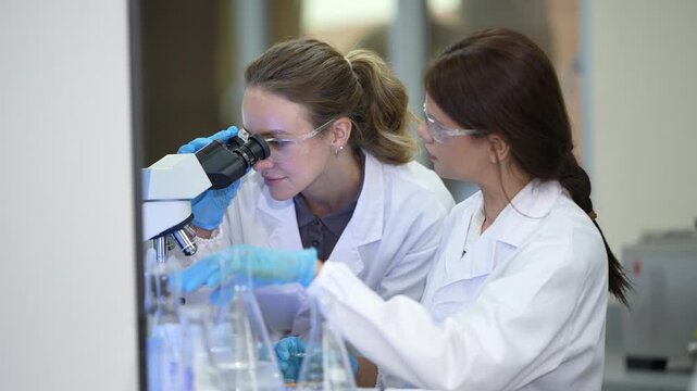 Two female medical researchers in white laboratory clothing, safety glasses and blue gloves lifting a vial past the microscope, then bending to the flasks below at a brightly lit laboratory.