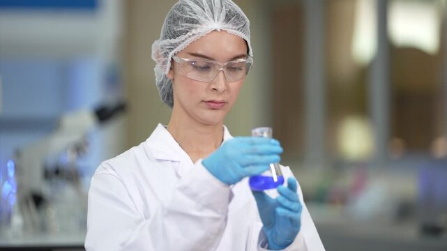 A female microbiologist in a white lab coat, hairnet and clear safety goggles swirling blue solution in a small flask, then lowering it out of frame at a brightly lit laboratory workbench.